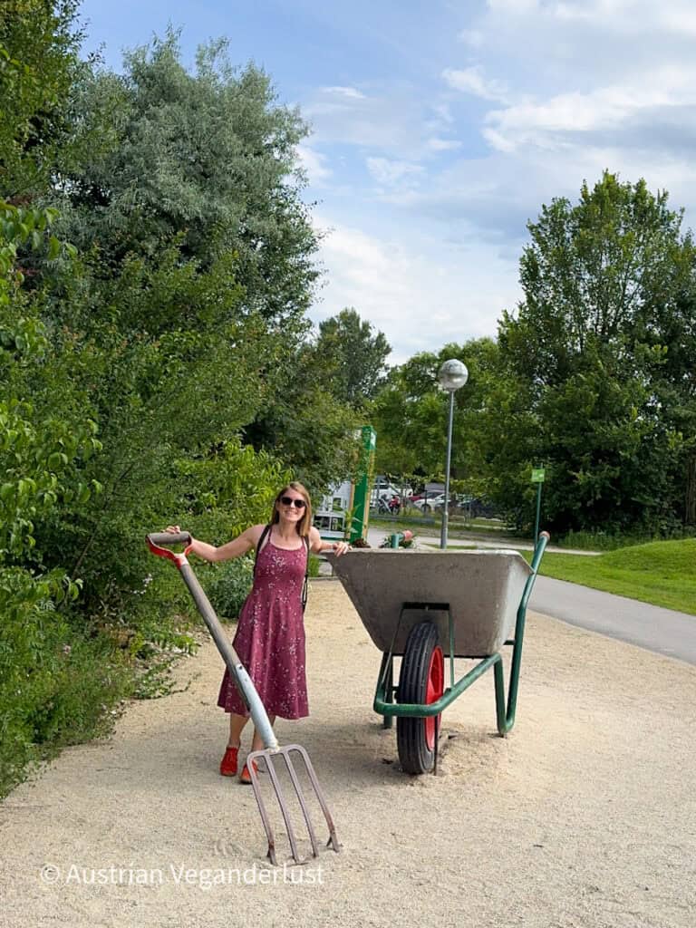 Tina in einem rosa Blumenkleid posiert neben überdimensionalen Skulpturen von Gartengeräten, darunter eine riesige Schubkarre und eine Heugabel. Die verspielte Installation steht an einem Kiesweg, umgeben von viel Grün.