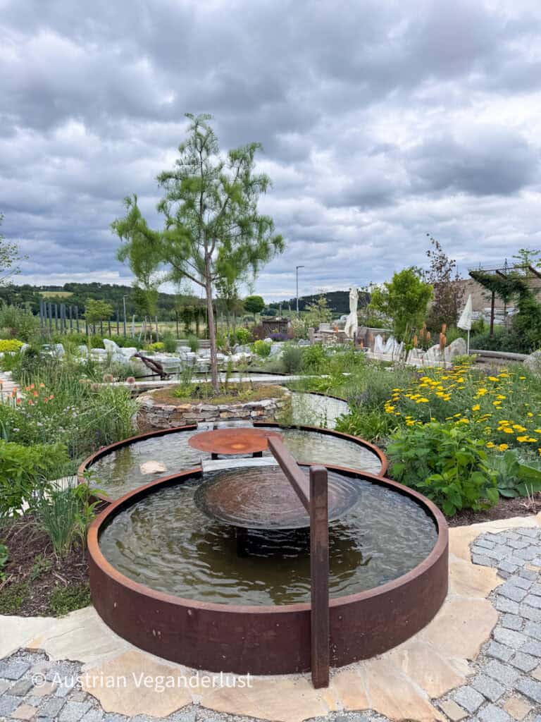 Ein rundes, rostfarbenes Wasserspiel sorgt in einem Schaugarten für plätschernde Spiegelungen zwischen Zierblumen, Gräsern und Steinwegen. Ein schlanker Baum ragt unter dramatischen Wolken aus der landschaftlich gestalteten Kulisse empor.