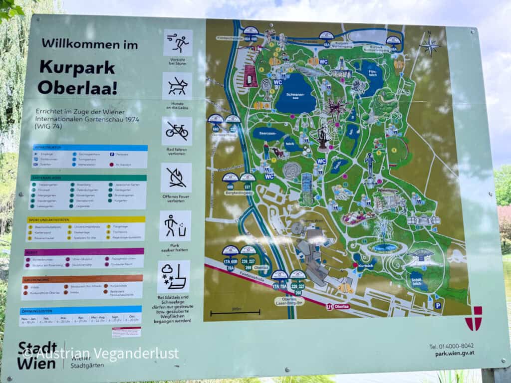 Information board at the entrance to Kurpark Oberlaa in Vienna showing a detailed illustrated map of the park with lakes, gardens, walking paths, attractions, and facilities. The sign includes the text "Willkommen im Kurpark Oberlaa!" along with visitor rules, opening hours, transportation information, and park amenities.