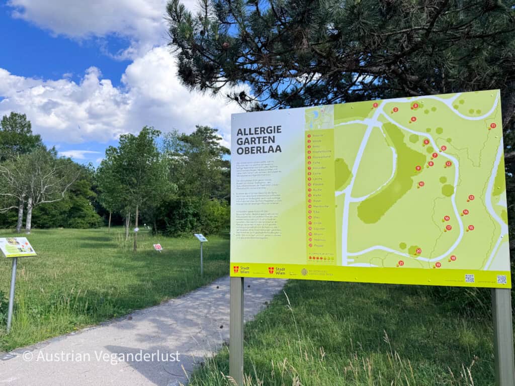 Sign for the Allergy Garden in Kurpark Oberlaa beside a walking path lined with trees and open grassland. The information board titled "Allergie Garten Oberlaa" displays a map and details about plant species selected to help visitors learn about common allergens.