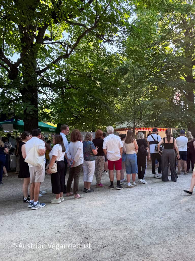 Eine lange Schlange von Konzertbesuchern wartet vor dem Sommernachtskonzert unter altem Baumbestand an den Essens- und Getränkeständen. Der schattige Kiesweg ist gesäumt von Besuchern, was die lange Wartezeit auf Erfrischungen vor der Vorstellung deutlich macht.