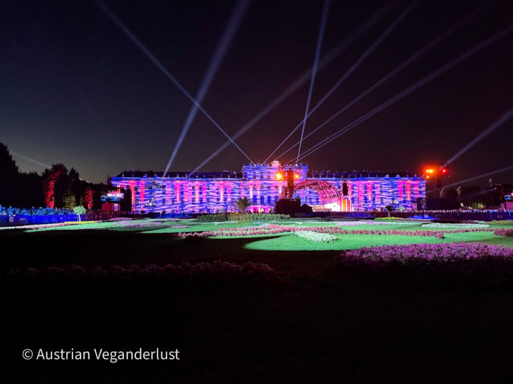 Das Schloss Schönbrunn wird während des Sommernachtskonzerts mit bunten Projektionen und weitreichenden Lichtstrahlen beleuchtet. Nach Sonnenuntergang erstrecken sich die formalen Blumengärten und die Konzertbühne über den Vordergrund.