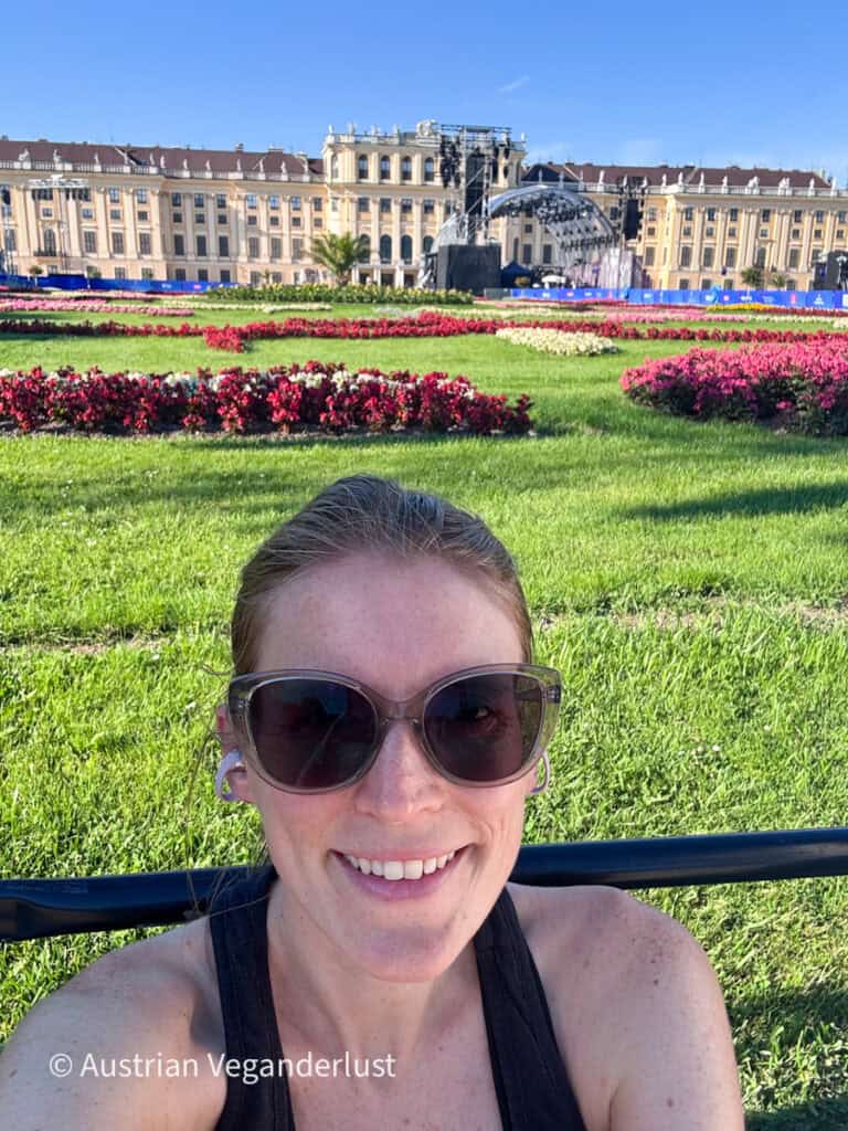 Tina, die eine Sonnenbrille trägt, posiert vor dem Sommernachtskonzert vor der Bühne und den Parterregärten des Schlosses Schönbrunn. Im Hintergrund sind an einem sonnigen Nachmittag die Konzertbühne und die Schlossfassade zu sehen.