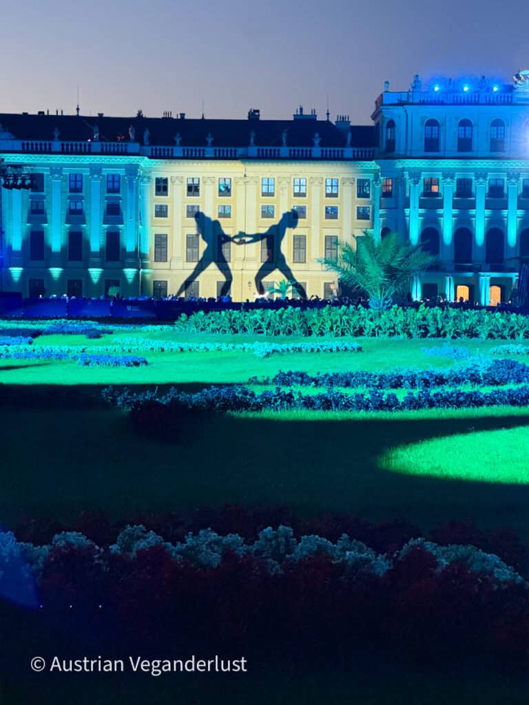 Das Schloss Schönbrunn ist während des Sommernachtskonzerts in blaues und grünes Licht getaucht, und die Silhouetten zweier Tänzer werden auf die Fassade projiziert. Die formalen Gärten im Vordergrund leuchten im bunten Schein der Bühnenbeleuchtung.
