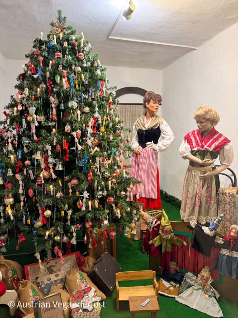 Decorated Christmas tree filled with vintage glass ornaments, candles, and handmade decorations stands beside mannequins dressed in traditional Austrian clothing. Antique toys, puppets, dolls, and children's furniture are arranged beneath the tree, recreating a nostalgic holiday display.