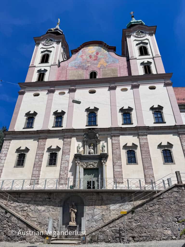 Tall Baroque church façade with twin clock towers and a pastel pink decorative pediment beneath a vivid blue sky. Stone steps lead up to the main entrance, which is framed by statues and carved architectural details.