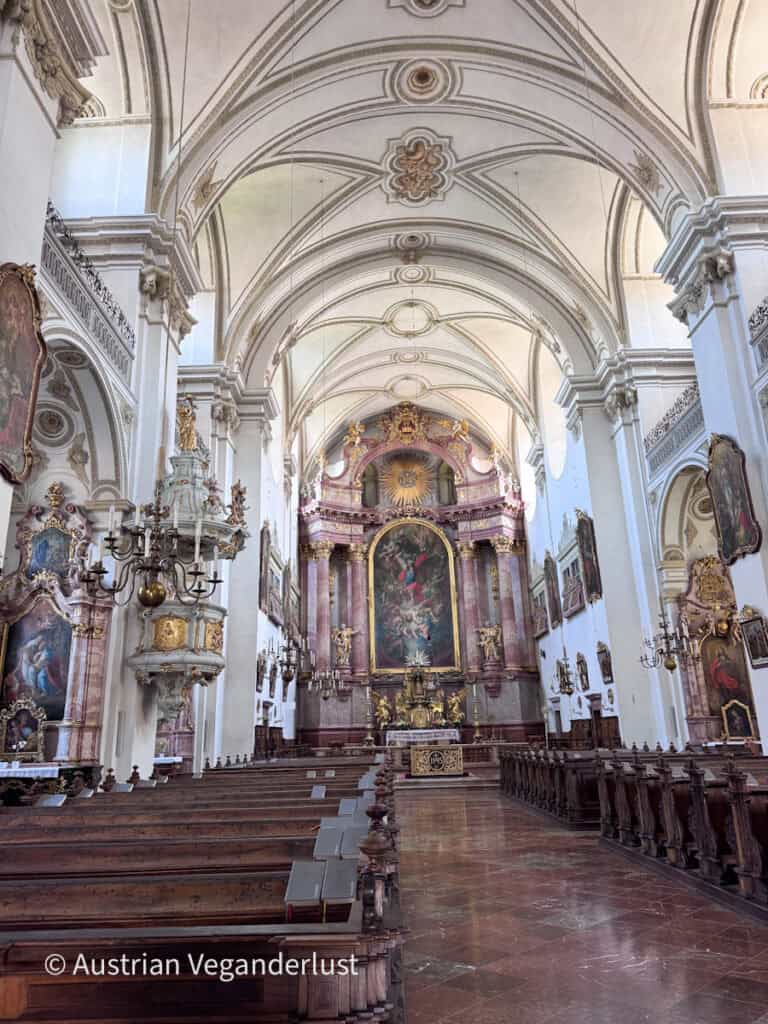 Ornate Baroque church interior with rows of wooden pews leading to a richly decorated high altar beneath a vaulted white ceiling. Marble columns, gilded details, and religious paintings line the spacious sanctuary.