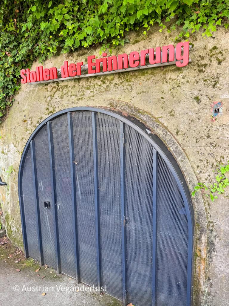 Arched tunnel entrance covered by a black metal gate built into a mossy stone wall with ivy growing above it. Red letters above the entrance read "Stollen der Erinnerung," marking the entrance to the memorial tunnel.