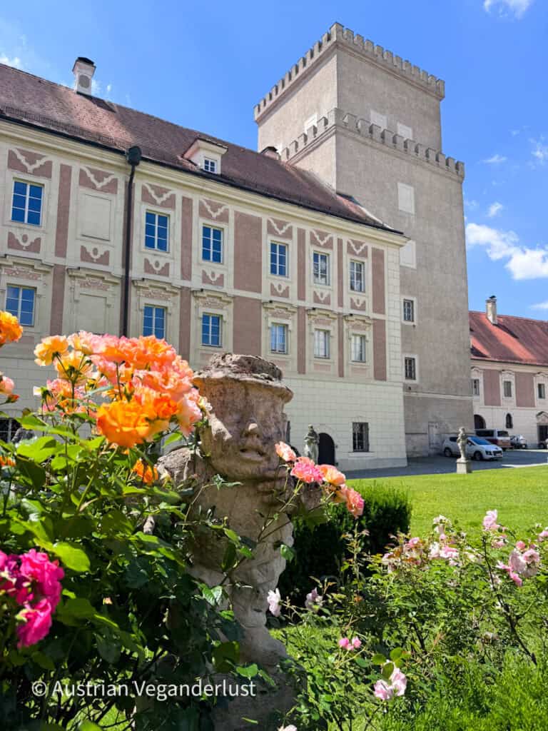 Formal castle garden filled with blooming orange and pink roses surrounds a carved stone sculpture. A historic palace building and square tower rise behind the flower beds on a sunny day.