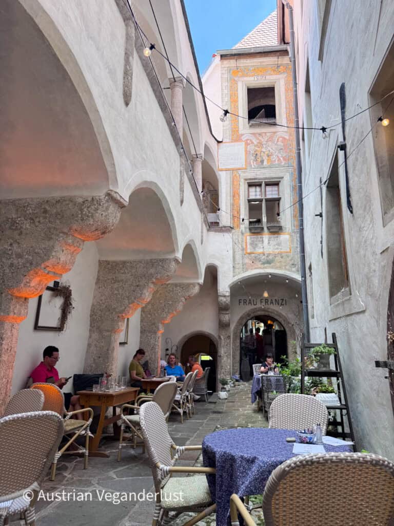 Narrow medieval courtyard lined with stone arches and café tables creates a peaceful place to dine in Steyr's old town. The entrance beneath the painted building is marked with the sign "FRÄU FRANZI."
