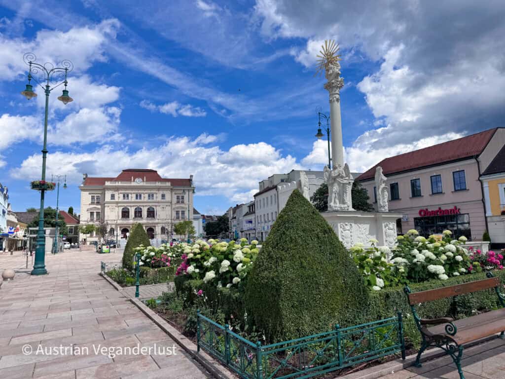 Der historische Hauptplatz in Tulln mit einer weißen Barock-Pest-Säule, umgeben von sorgfältig geschnittenen Hecken und blühenden Hortensien. Verzierte Straßenlaternen und elegante Stadtgebäude säumen den Fußgängerplatz unter einem strahlend blauen Himmel mit vereinzelten Wolken.