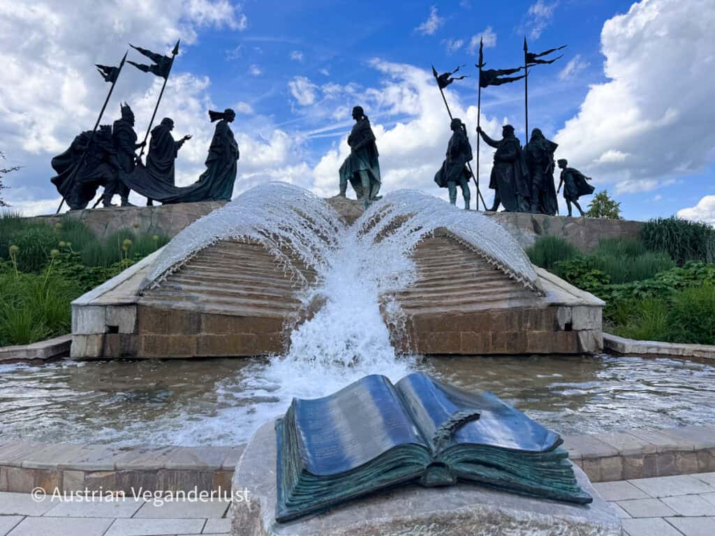 Ein Springbrunnen-Denkmal mit historischen Figuren aus Bronze, die Fahnen tragen und sich über ein herabfließendes Wasser hinweg gegenüberstehen. Im Vordergrund steht eine offene Buchskulptur aus Bronze, während das Wasser unter einem dramatischen, wolkenverhangenen Himmel über Steinstufen hinwegspritzt.