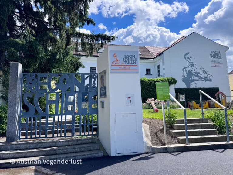 Der Eingang zum Egon-Schiele-Museum in Tulln mit einem verzierten Metalltor, auf dem der Name des Künstlers steht, und einem großen Wandporträt an der Gebäudefassade. Museumsbeschilderung, Gartenbepflanzung und eine kleine Treppe führen die Besucher in das ehemalige Geburtshaus und Museum, das Egon Schiele gewidmet ist.