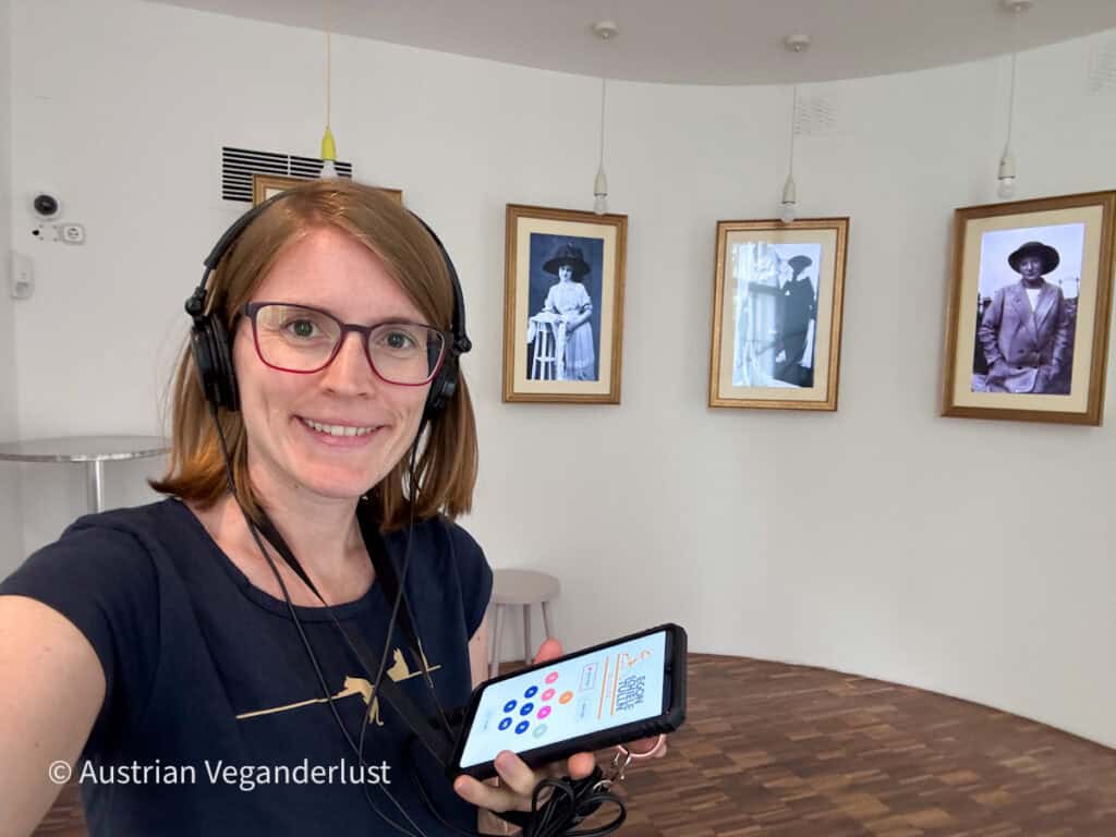 Tina trägt Kopfhörer und hält einen Audioguide im Egon-Schiele-Museum in Tulln in der Hand. Hinter ihr hängen schwarz-weiße Porträtfotos an der geschwungenen Galeriewand, während sie sich den Ausstellungskommentar anhört.