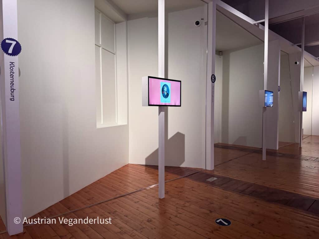 Interaktiver Ausstellungsraum im Egon-Schiele-Museum mit nummerierten Stationen und digitalen Bildschirmen, auf denen historische Inhalte gezeigt werden. Weiße Trennwände und polierte Holzböden sorgen für eine ruhige, moderne Museumsatmosphäre.