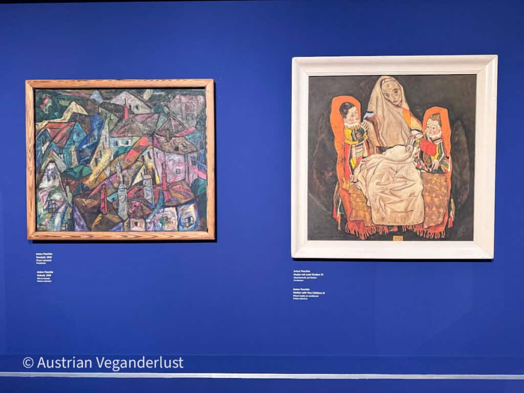 Zwei Gemälde, die an einer tiefblauen Museumswand im Egon-Schiele-Museum ausgestellt sind. Das eine Bild zeigt eine abstrakte Stadtlandschaft, das andere eine sitzende Mutter mit zwei Kindern im ausdrucksstarken Stil des frühen 20. Jahrhunderts.