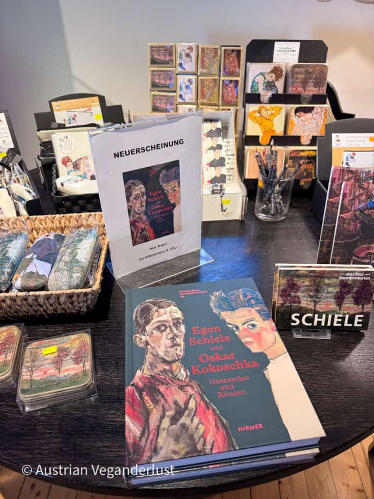 Auslage im Museumsshop mit Büchern, Postkarten, Schreibwaren und Souvenirs, die von Egon Schieles Kunstwerken inspiriert sind. Im Vordergrund ist ein Buch mit dem Titel „Egon Schiele und Oskar Kokoschka. Netzwerker und Rivalen“ prominent ausgestellt.