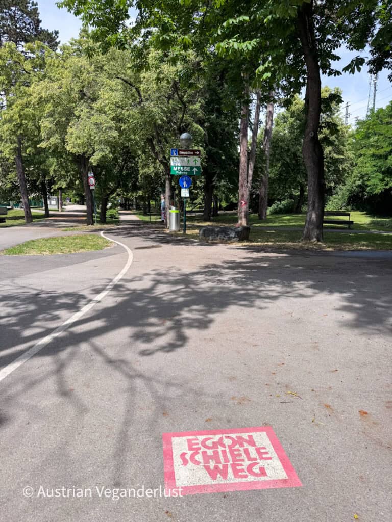 Rosa Wegweiser mit der Aufschrift „Egon Schiele Weg“, aufgemalt auf einem gepflasterten Weg unter schattigen Bäumen in Tulln. Wegweiser und Wanderwege zeigen einen Teil der Route, die Orte verbindet, die mit dem Künstler in Verbindung stehen.
