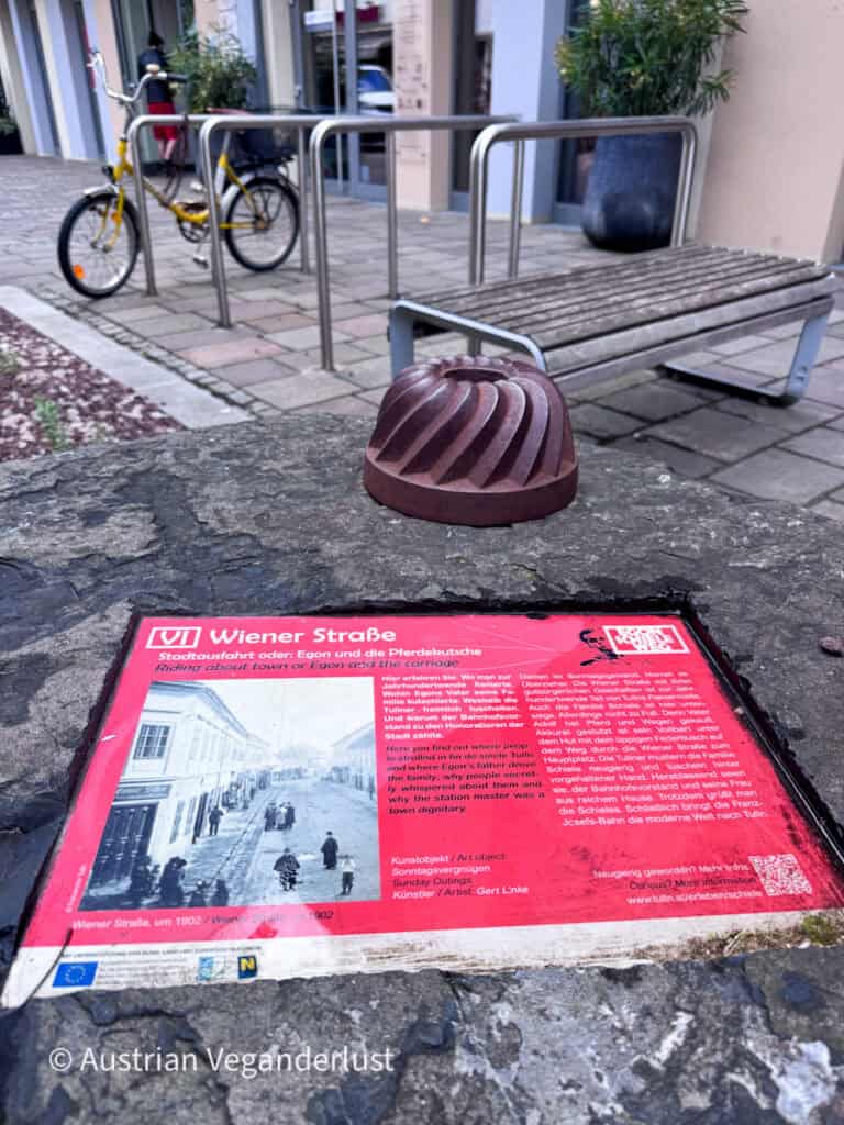 Eine Informationstafel zum Egon-Schiele-Weg, angebracht auf einem Steinsockel an der Wiener Straße in Tulln. Historische Fotos und ein Text erläutern den Zusammenhang zwischen der Straße, dem frühen Leben des Künstlers und der Geschichte der Stadt.