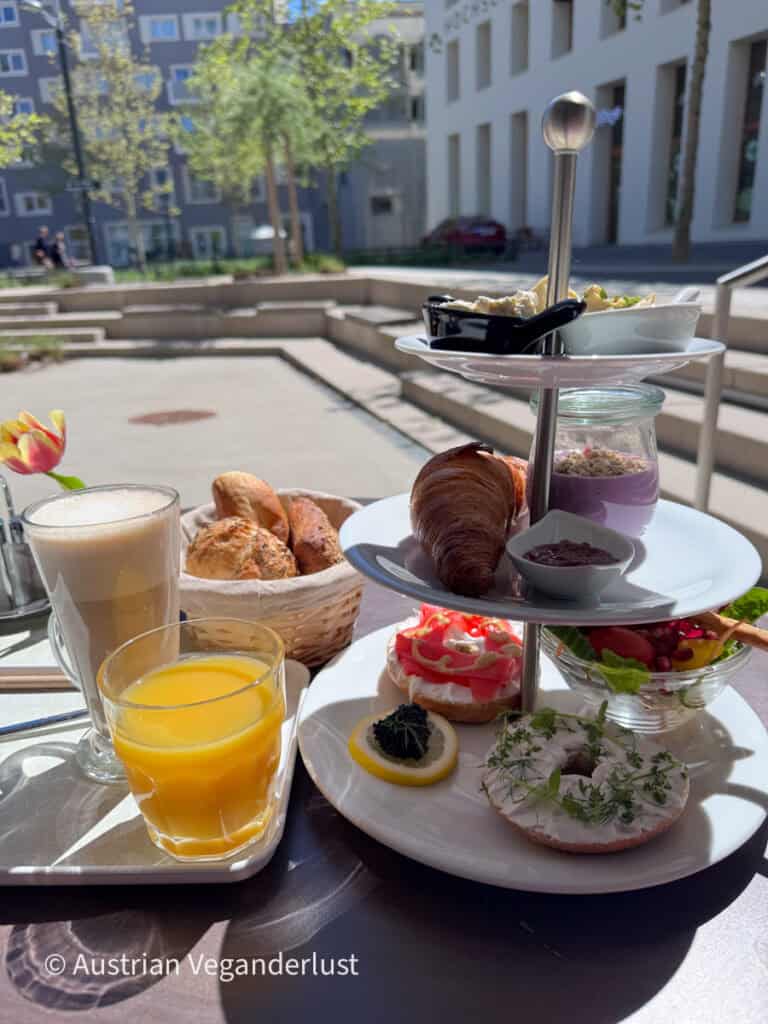 Ein dreistöckiges Frühstücksset steht auf einem Café-Tisch im Freien, bestückt mit Gebäck, Bagels mit Frischkäse und Kräutern, Joghurt, Salat und kleinen Schälchen mit Aufstrichen. Daneben stehen ein Glas Orangensaft und ein Latte, im Hintergrund ist ein sonniger Innenhof zu sehen.