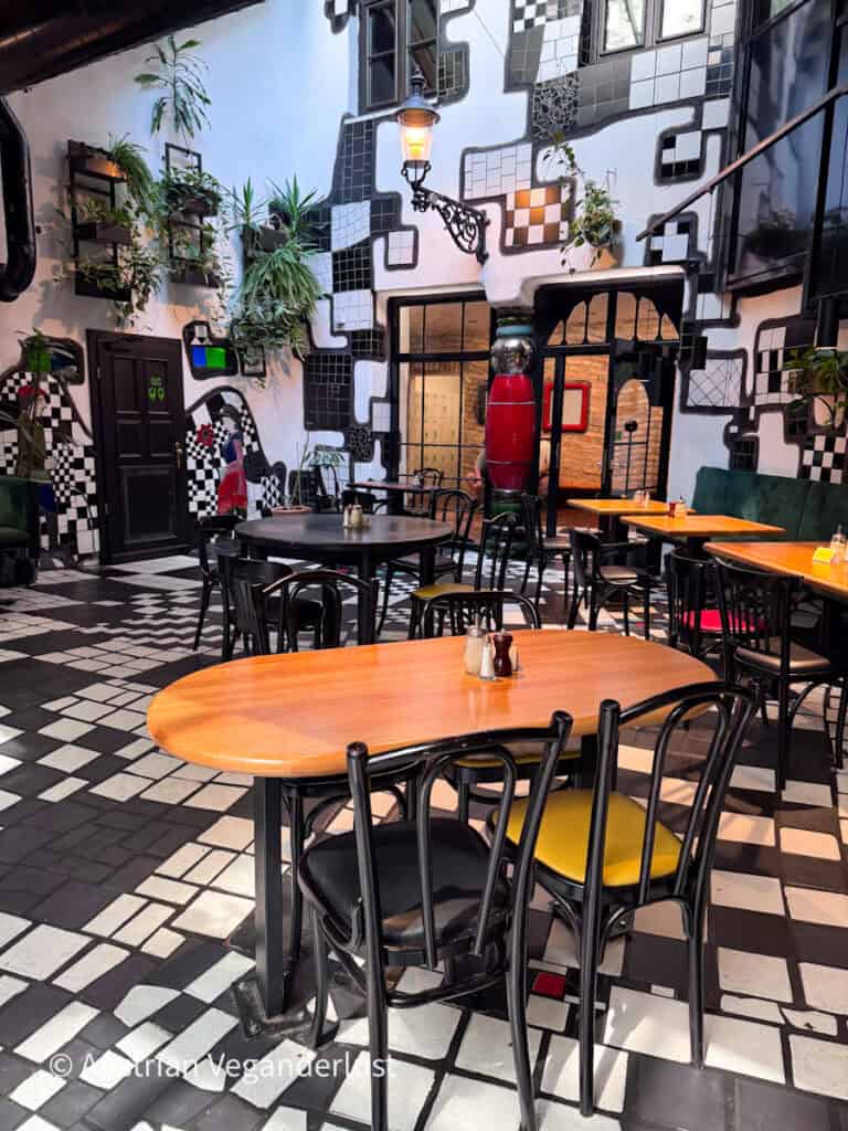 Interior dining area with black and white tiled floors and walls designed in an irregular pattern. Wooden tables, black chairs, hanging plants, and decorative columns create a whimsical atmosphere.