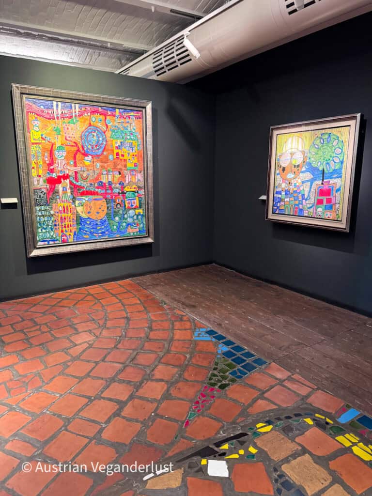 Gallery room displaying two brightly colored paintings on dark walls. A curved brick floor with mosaic tile inlays reflects the organic design style associated with Friedensreich Hundertwasser.