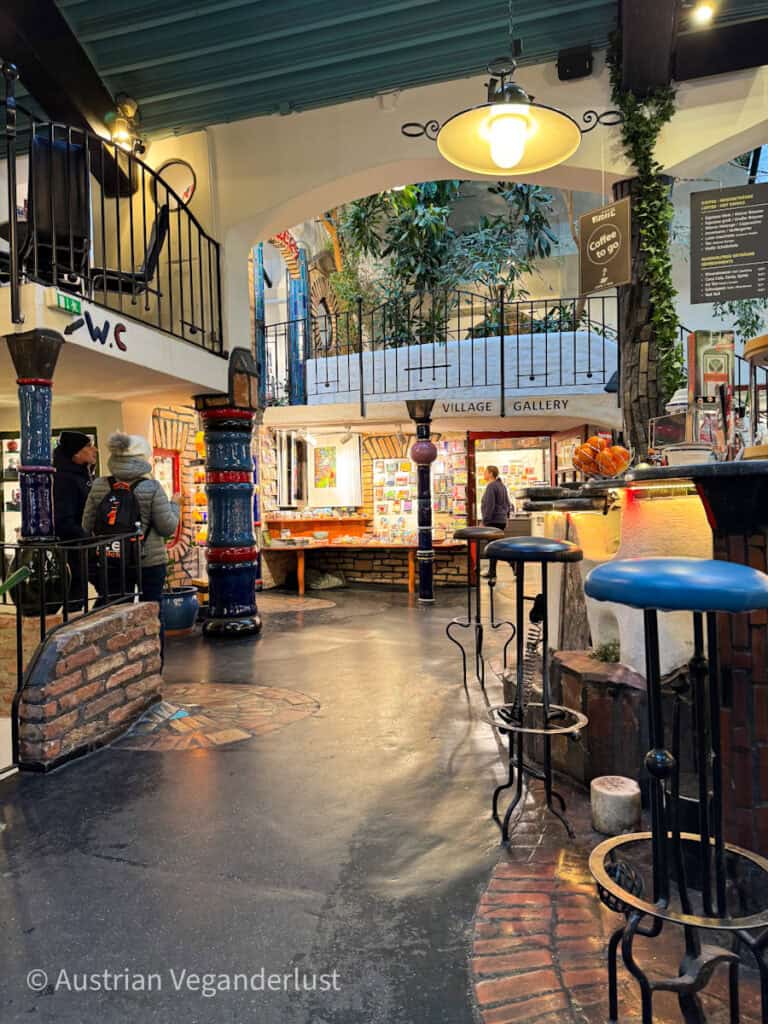 Interior hall with colorful columns, a café counter, and an art gallery space. Signs reading "Village Gallery" and "Coffee to go" hang above walkways lined with plants and decorative architectural details.