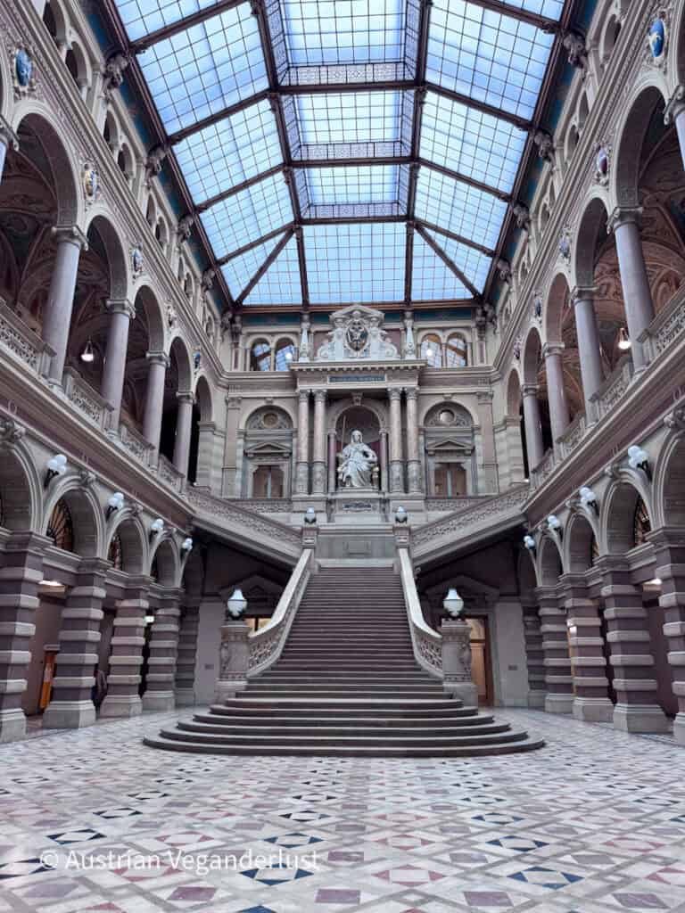 Weitwinkelaufnahme einer prächtigen zentralen Treppe, die zu einer sitzenden Statue in einem riesigen, von einem Glasdach überdachten Atrium hinaufführt. Symmetrische Bögen, Marmorsäulen, dekorative Schnitzereien und gemusterte Steinböden unterstreichen die Pracht des historischen Innenraums.