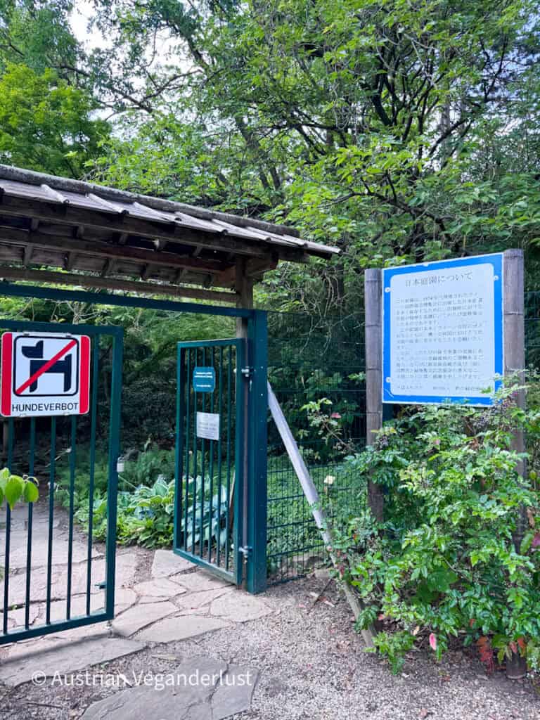 Eingangstor zu einem japanischen Garten mit einem Holzdach und viel Grün drumherum. Neben einer Informationstafel mit japanischem Text hängt ein Schild mit der Aufschrift „HUNDEVERBOT“.