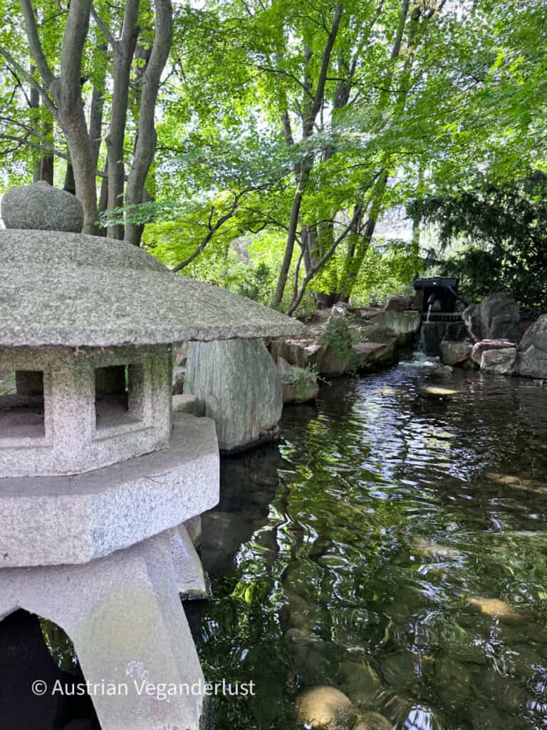 Neben einem klaren Teich, der von Felsbrocken und Bäumen gesäumt ist, steht eine traditionelle Steinlaterne. Im Hintergrund speist ein kleiner Wasserfall das Wasser, in dem sich das umliegende Grün spiegelt.