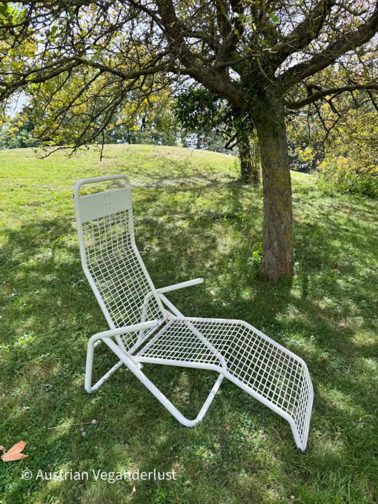 A white metal reclining chair sits beneath a large shady tree on a grassy hillside. Dappled sunlight filters through the branches, creating a quiet spot for relaxing in the gardens.