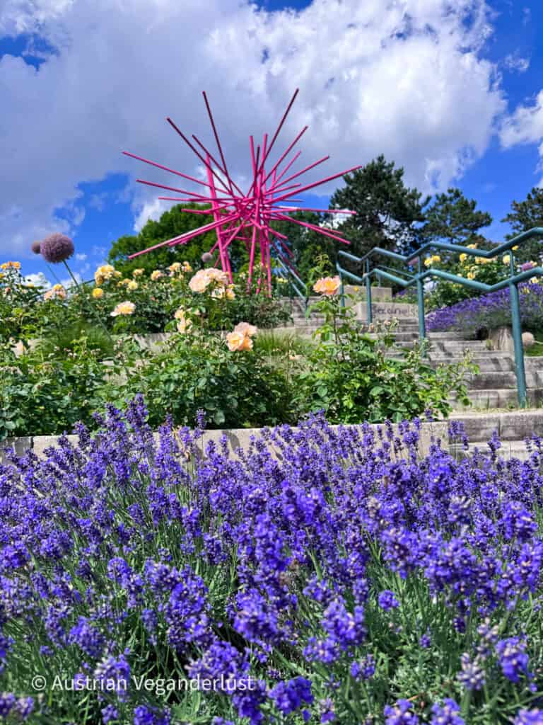 Im Vordergrund eines terrassierten Rosengartens mit Steinstufen und Geländern blühen violette Lavendelblüten. Vor einem blauen, wolkenverhangenen Himmel ragt eine leuchtend rosa abstrakte Skulptur über die Blumenbeete hinaus.