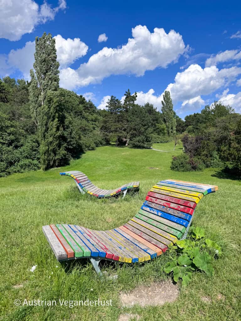 Two colorful curved wooden lounge benches sit in a grassy meadow surrounded by trees and open green space. The painted slats in red, blue, yellow and green add a playful contrast to the natural landscape.