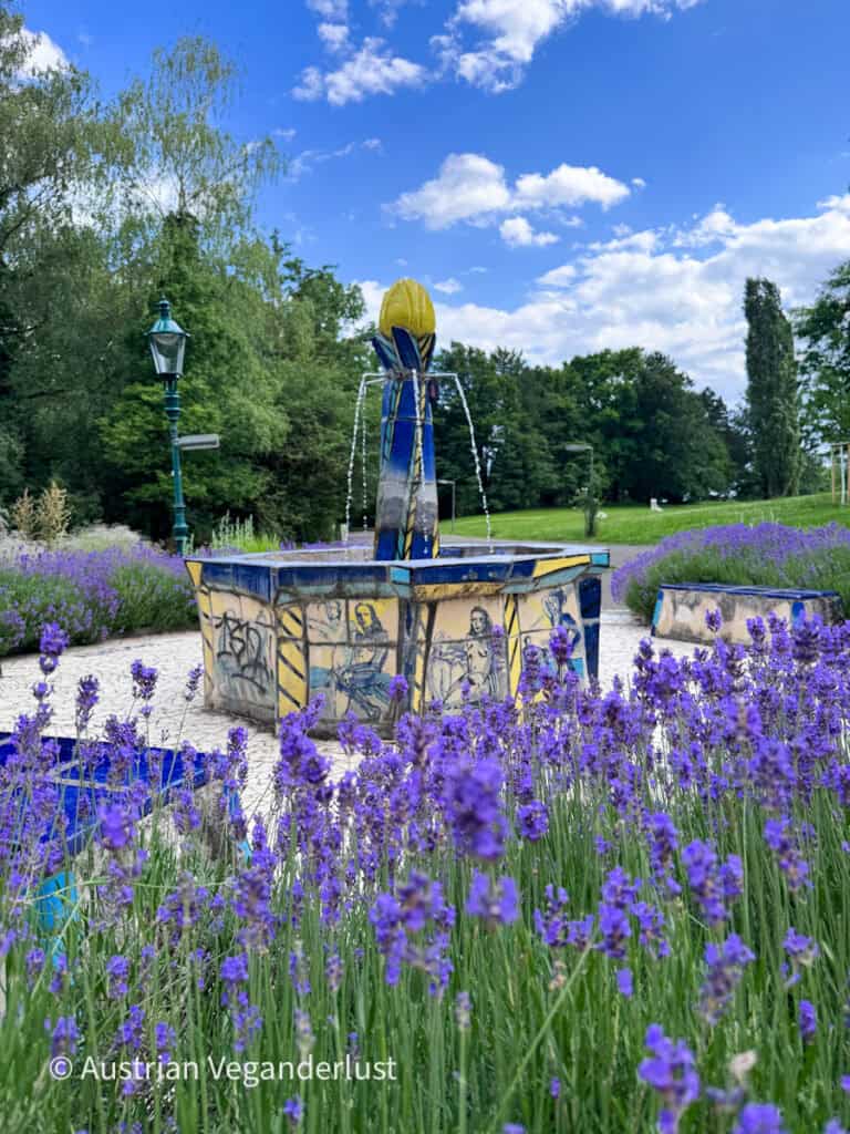 A decorative tiled fountain surrounded by blooming purple lavender stands in a landscaped garden. Water streams from a yellow flower shaped sculpture at the center beneath a bright blue sky with scattered clouds.