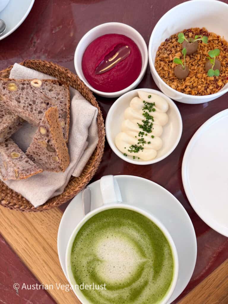 Draufsicht auf ein veganes Frühstücksbuffet mit einem schaumigen grünen Matcha-Latte, Scheiben Körnerbrot, cremigen pflanzlichen Aufstrichen mit Schnittlauch, leuchtend rosa Rote-Bete-Hummus und herzhaften Häppchen, die in kleinen Schälchen auf einem Holztablett angerichtet sind.
