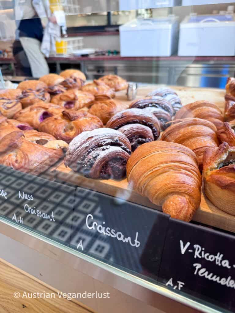 Glasvitrine in einer Bäckerei mit goldbraunen Croissants, Schokoladengebäck und fruchtgefüllten Plunderteilchen. Auf den handgeschriebenen Schildern stehen unter anderem „Croissant“, „Nuss Croissant“ und „V-Ricotta“.