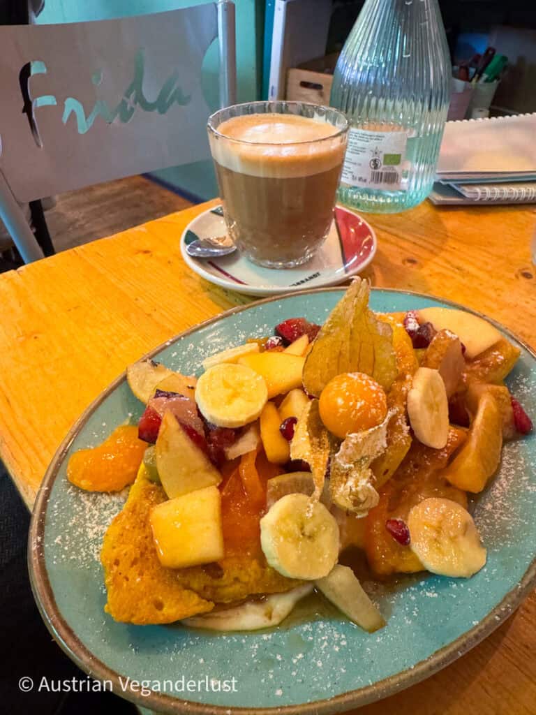 Dicke Scheiben French Toast, garniert mit Banane, Apfel, Beeren, Zitrusfruchtstücken und Puderzucker, auf einem türkisfarbenen Teller. Hinter dem mit Obst belegten Frühstücksteller steht ein Latte in einer Glastasse auf einem gemütlichen Café-Tisch.