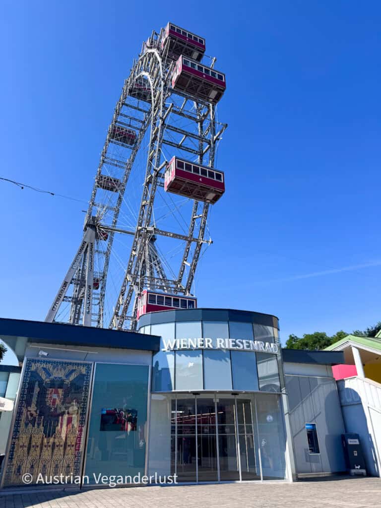 Das Eingangsgebäude des Wiener Riesenrads mit dem Schriftzug „WIENER RIESENRAD“ über den Glastüren. Das historische Riesenrad ragt hoch über einem empor, und vor dem strahlend blauen Himmel sind mehrere rote Fahrgastkabinen zu sehen.