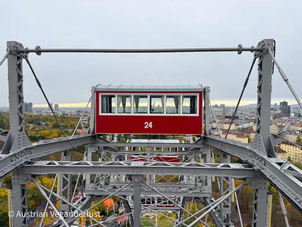 Die rote Riesenrad-Gondel mit der Nummer 24 hängt im Stahlgerüst des Wiener Riesenrads. Die Gondel schwebt hoch über Wien, und in der Ferne sind unter einem bewölkten Himmel die Gebäude der Stadt und herbstlich gefärbte Bäume zu sehen.