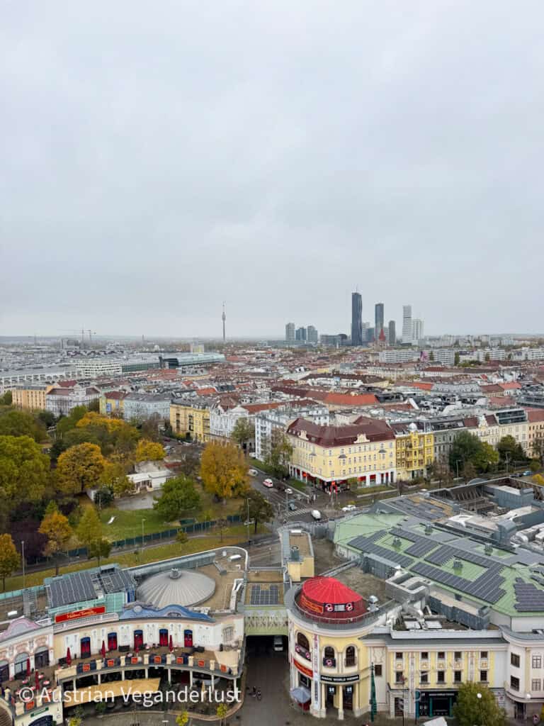 Blick über Wien vom Wiener Riesenrad aus, der Reihen historischer Gebäude, von von Bäumen gesäumten Straßen und modernen Wolkenkratzern am Horizont zeigt. Tiefe graue Wolken hängen über der Stadt.