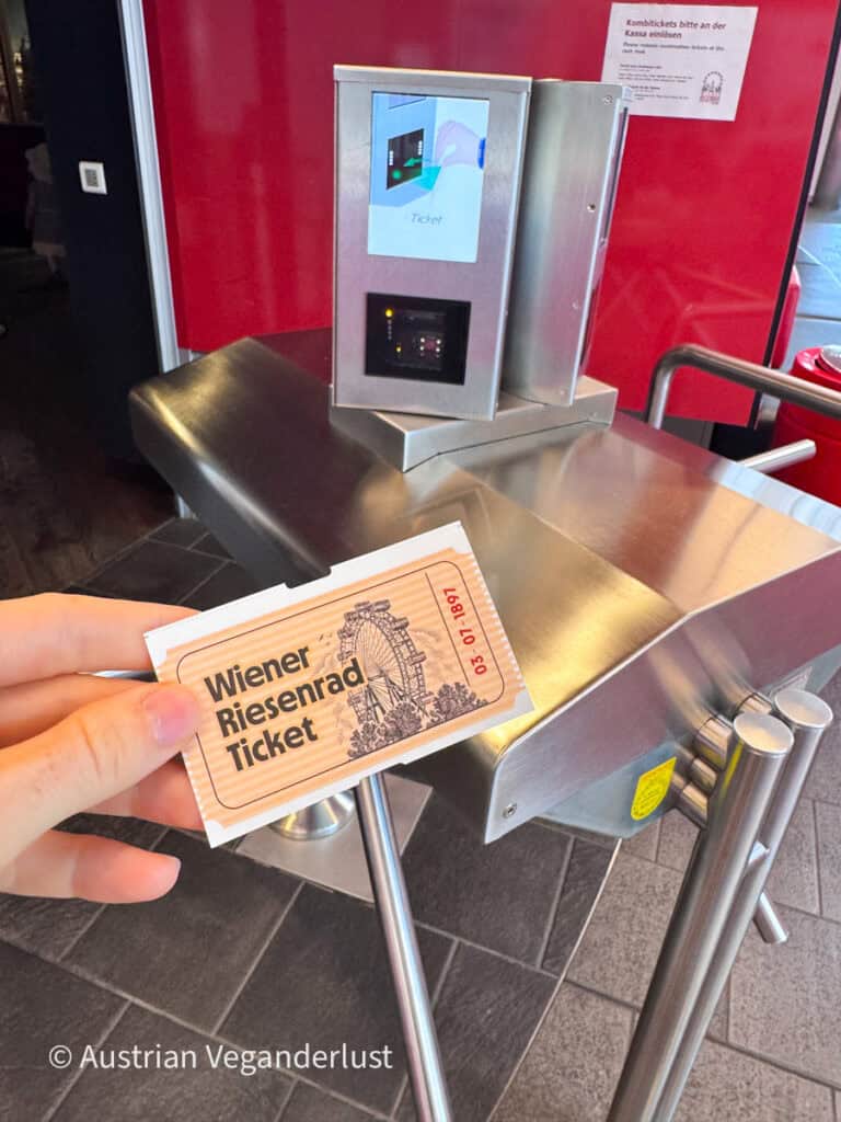 Eine Hand, die eine Eintrittskarte für das Wiener Riesenrad hält, neben einem Drehkreuz und einem Ticketscanner. Auf der Karte steht „Wiener Riesenrad Ticket“.