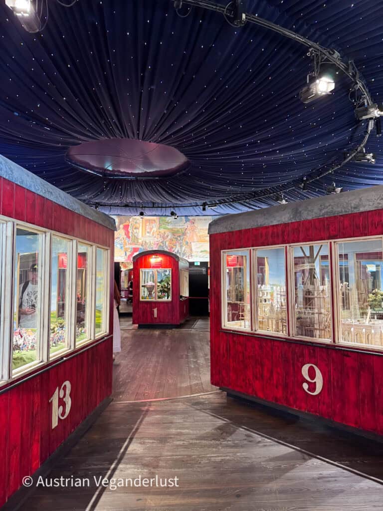 Ausstellungshalle im Inneren des Wiener Riesenrads mit roten, nummerierten Aussichtsgondeln, die entlang eines Holzstegs angeordnet sind. Eine dunkelblaue Decke, die mit kleinen Lichtern übersät ist, sorgt für einen sternenklaren Effekt über den Köpfen.