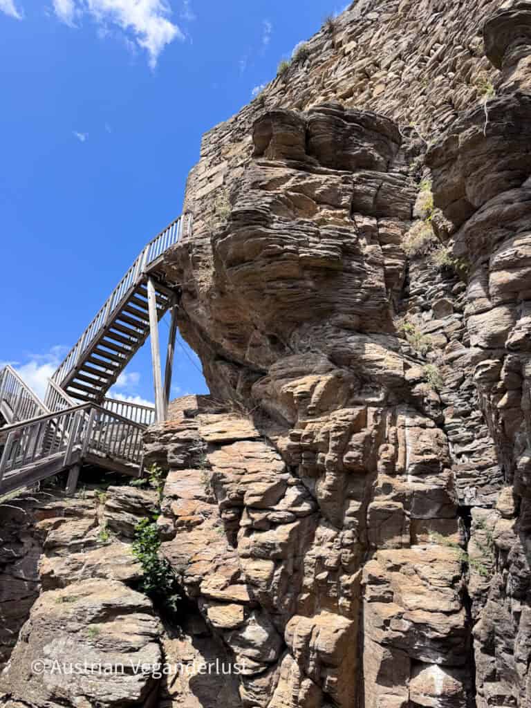 Massive rock face supporting the walls of Burgruine Aggstein with a wooden staircase climbing to an elevated viewing platform. The castle is built directly into the rugged cliffs beneath a clear blue sky.