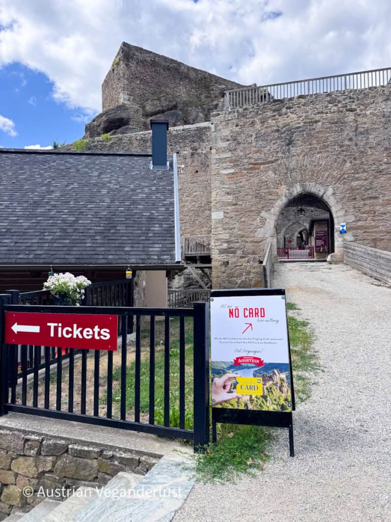 Entrance to Burgruine Aggstein with a stone gateway, ticket office and signs directing visitors. The red sign reads "Tickets" while the white sign says "Mit der NÖ Card hier entlang" and "NO CARD hier entlang" with an arrow pointing towards the entrance.