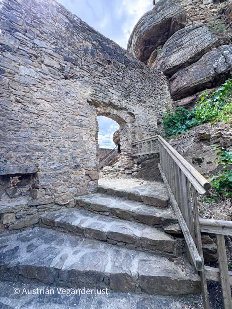 Stone steps and a wooden handrail lead through an arched gateway built into the medieval walls of Burgruine Aggstein. Massive rock formations rise above the castle walls beside the entrance.