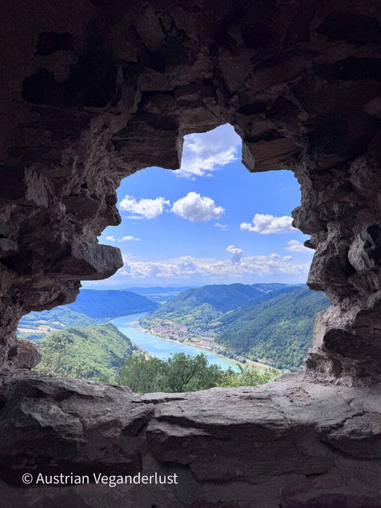View of the Danube River and the Wachau Valley framed by a rough stone window opening inside Burgruine Aggstein. Blue sky with scattered clouds stretches above the river and surrounding forested hills.