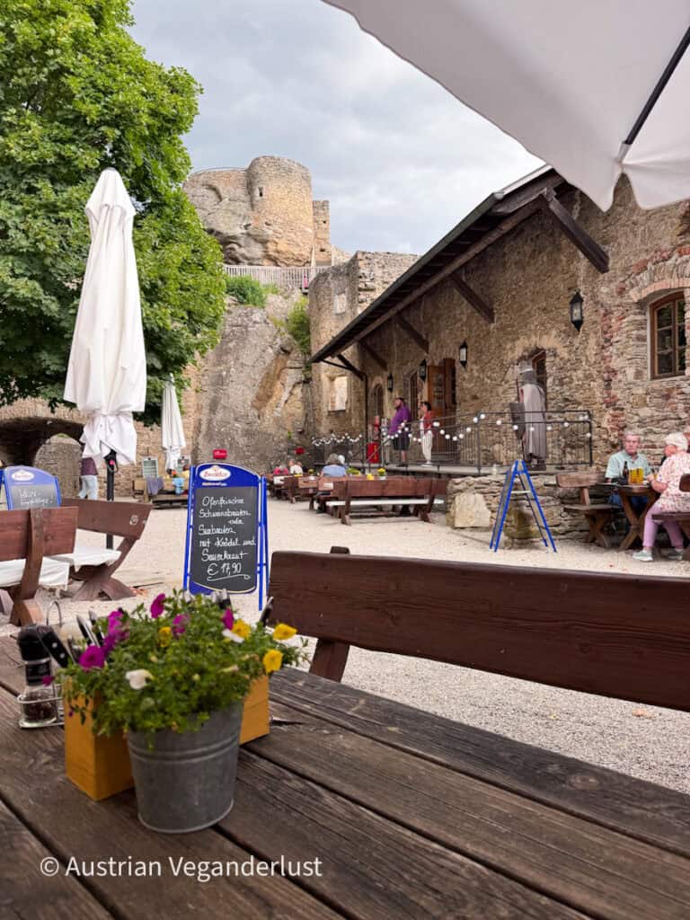 Outdoor courtyard restaurant at Burgruine Aggstein with wooden picnic tables, flower planters and medieval stone buildings. A chalkboard sign reads "Ofenfrischer Schweinsbraten oder Surbraten mit Knödel und Sauerkraut € 17,90" while visitors enjoy meals beneath the castle walls.