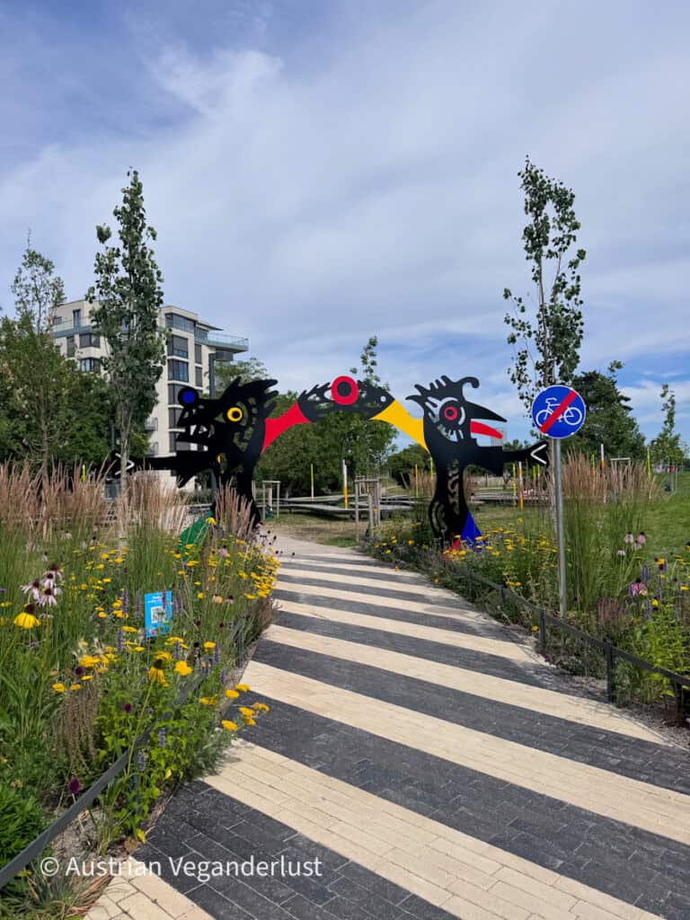 Eine farbenfrohe, abstrakte Skulptur überspannt einen Fußweg durch einen landschaftlich gestalteten Park voller Wildblumen und Ziergräser. Die schwarze Skulptur besticht durch kräftige rote, gelbe, blaue und weiße Akzente und bildet so einen auffälligen Blickfang vor dem Hintergrund des umgebenden Grüns.