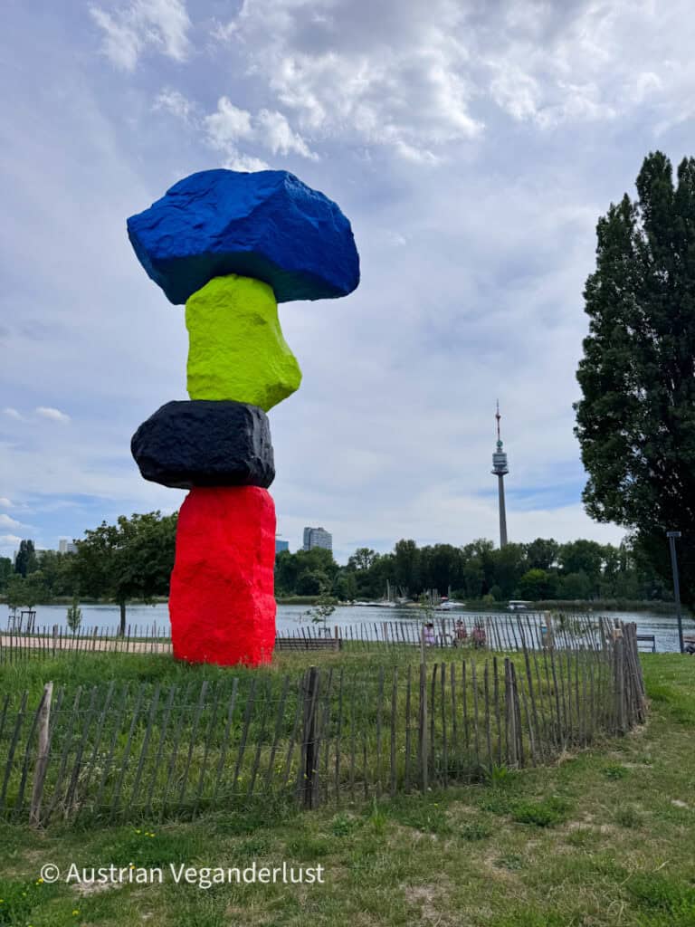 Eine farbenfroh bemalte Skulptur aus aufeinandergestapelten Felsbrocken in Blau, Limettengrün, Schwarz und Rot steht neben einem Park am Seeufer mit Bäumen und der Wiener Skyline im Hintergrund. Hinter dem Wasser ragt unter einem dramatischen Wolkenhimmel ein hoher Aussichtsturm empor.