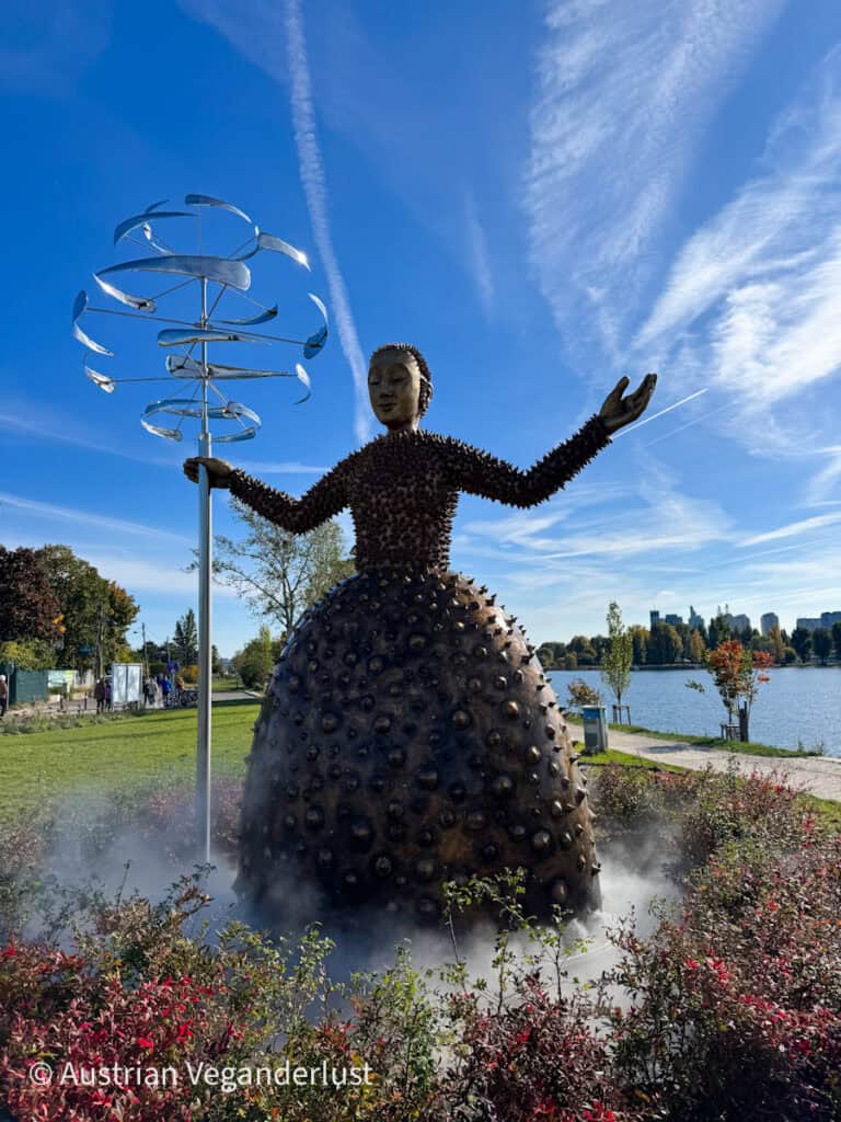 Eine große Bronzeskulptur einer Frau mit ausgestreckten Armen steht neben einer kinetischen Windskulptur aus Metall, die auf einen See blickt. Leichter Nebel umgibt den Sockel des Kunstwerks, während ein strahlend blauer Himmel mit zarten Wolkenfäden den modernen Skulpturengarten einrahmt.
